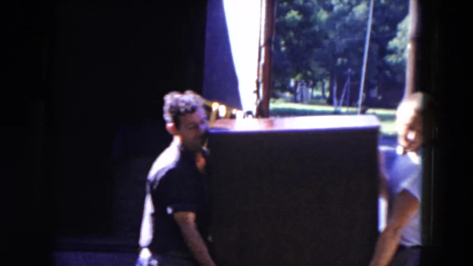 Adult men carry a large cabinet or furniture piece near an open doorway, with daylight and trees visible beyond.