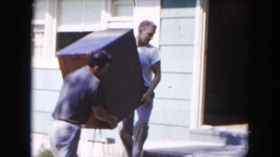 Two adult men handle a heavy furniture piece outside a light-colored Hicksville house in 1954 home-movie footage.