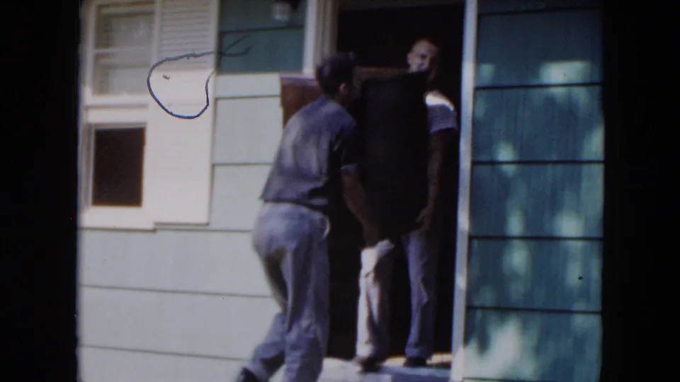 Adult men maneuver a large furniture piece by a doorway and green siding, consistent with the 1954 Hicksville moving scene.
