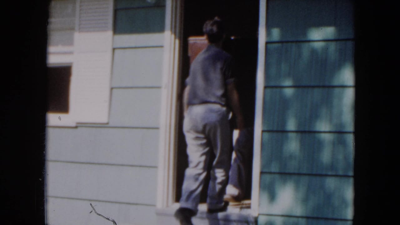 In 1954 Hicksville home-movie footage, a man stands at a residential doorway beside blue siding and a light exterior wall.