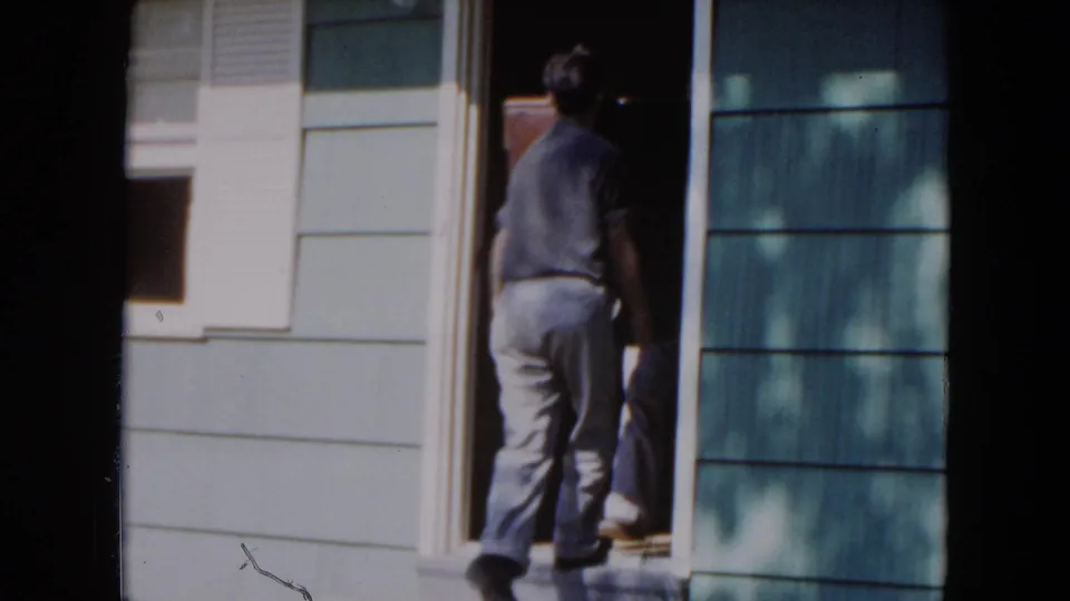 In 1954 Hicksville home-movie footage, a man stands at a residential doorway beside blue siding and a light exterior wall.