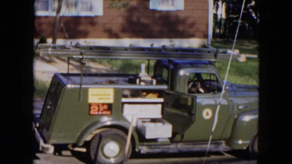 A green mid-century utility truck with roof ladder and rear equipment is parked on a Hicksville residential street.