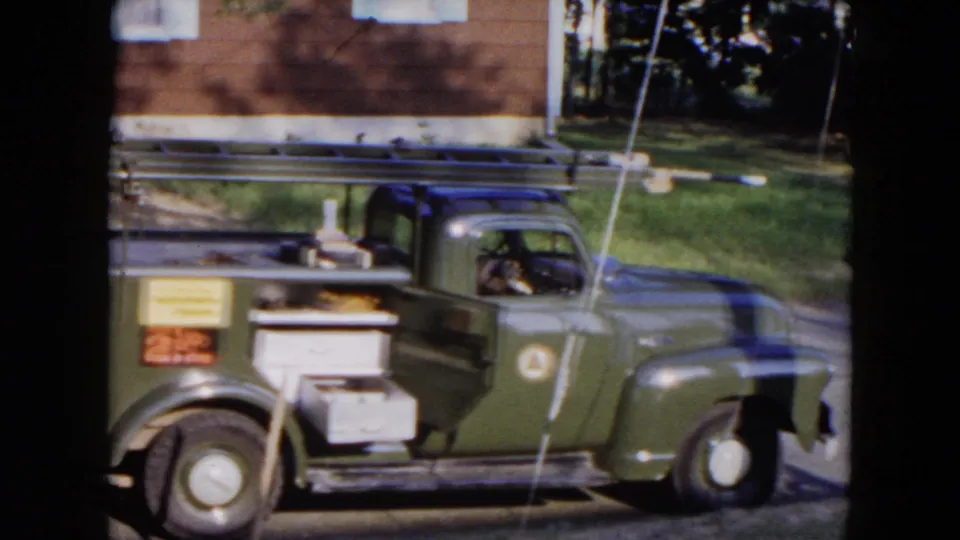 The 8mm frame shows a green 1950s utility truck in side view, parked by grass and a brick building in Hicksville.