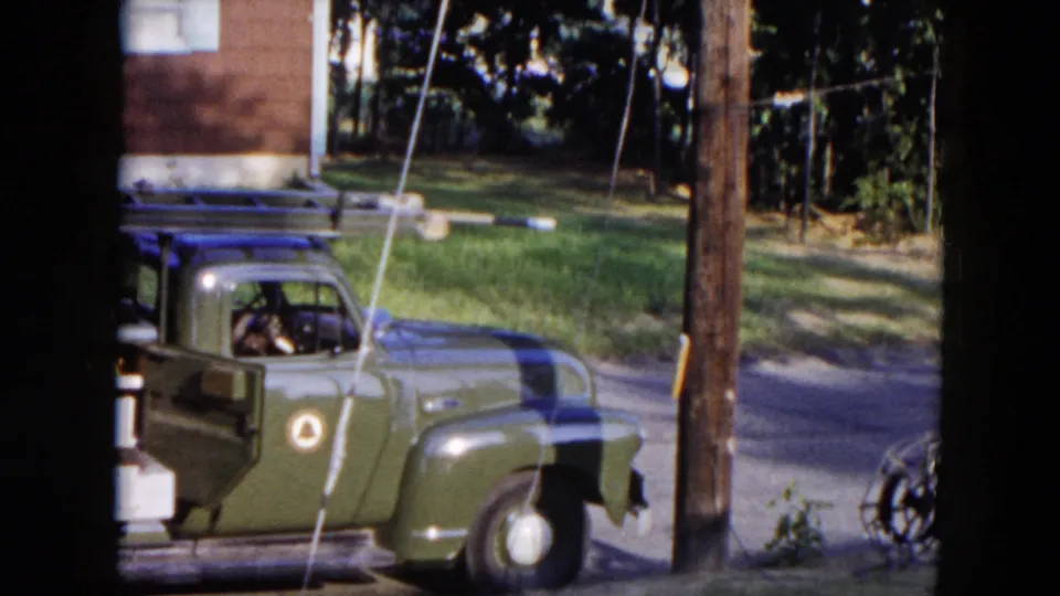 A green utility truck is partly framed beside a wooden utility pole, with lawn and trees beyond the sunny residential street.