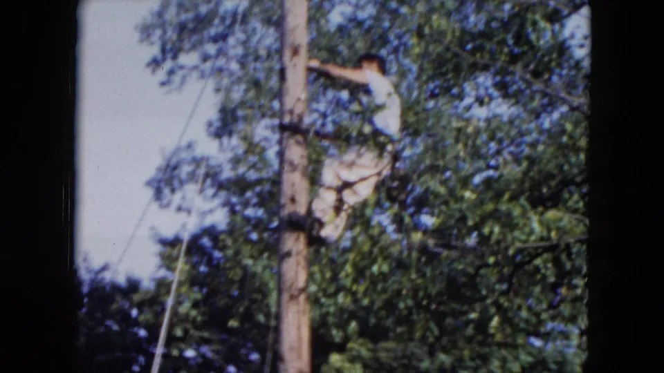 1954 Hicksville home-movie frame of the utility worker leaning from the wooden pole as he reaches toward the line or hardware.