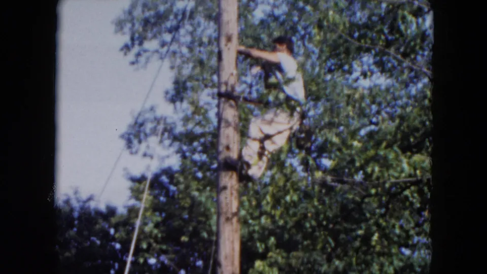 In the Hicksville 1954 home-movie frame, a man appears beside a utility pole with trees and open sky around him.