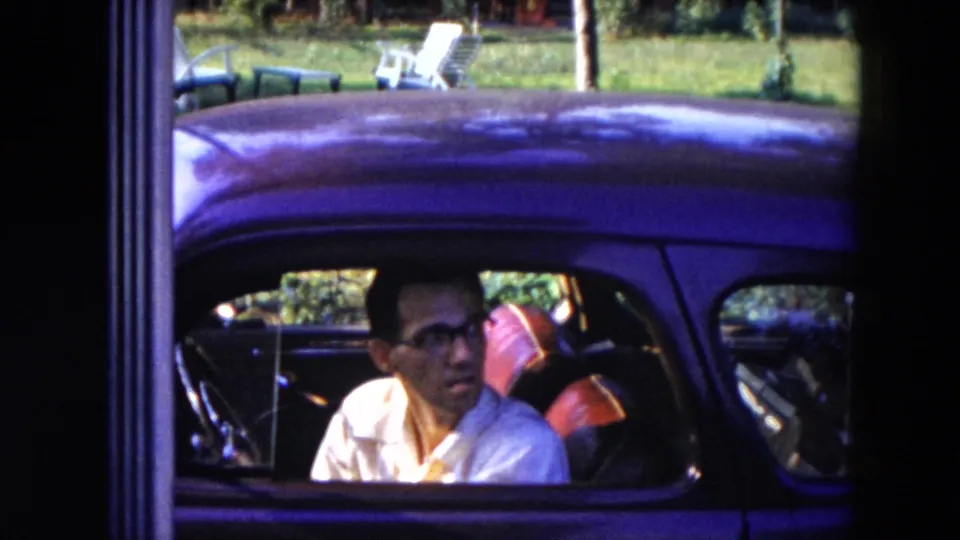 A man in a white shirt and glasses sits inside a dark rounded car in the Hicksville home-movie scene.