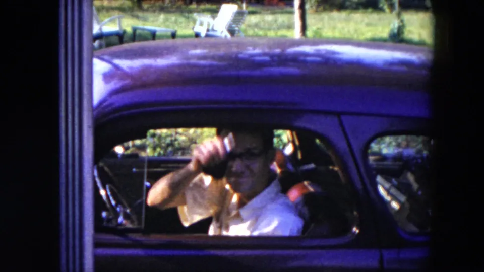 The man raises a hand toward the camera from inside the dark car, with red seats and grass visible behind him.