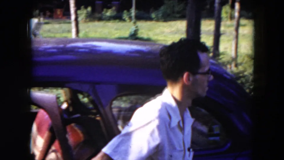 The man has exited or is moving beside the dark car in the 1954 Hicksville outdoor home-movie frame.