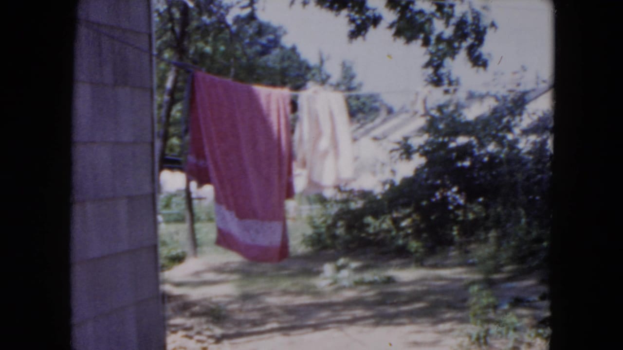 1954 Hicksville backyard view with laundry on a clothesline, including a pink towel, near a house wall and trees.
