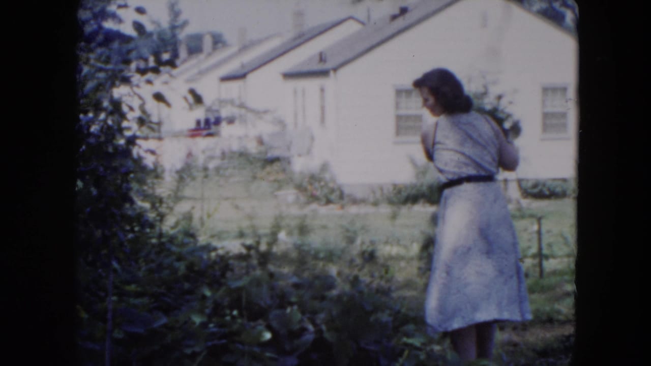 Woman in a light dress stands in a backyard near leafy plants and a white house.