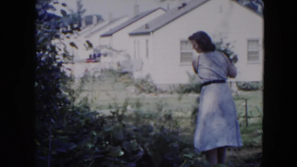 In Hicksville, New York, a woman in a light dress stands beside overgrown backyard foliage with a white house behind her.