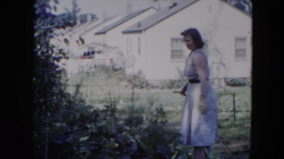 The woman stands beside the garden edge and narrow yard path, with the Hicksville residential exterior still visible behind her.