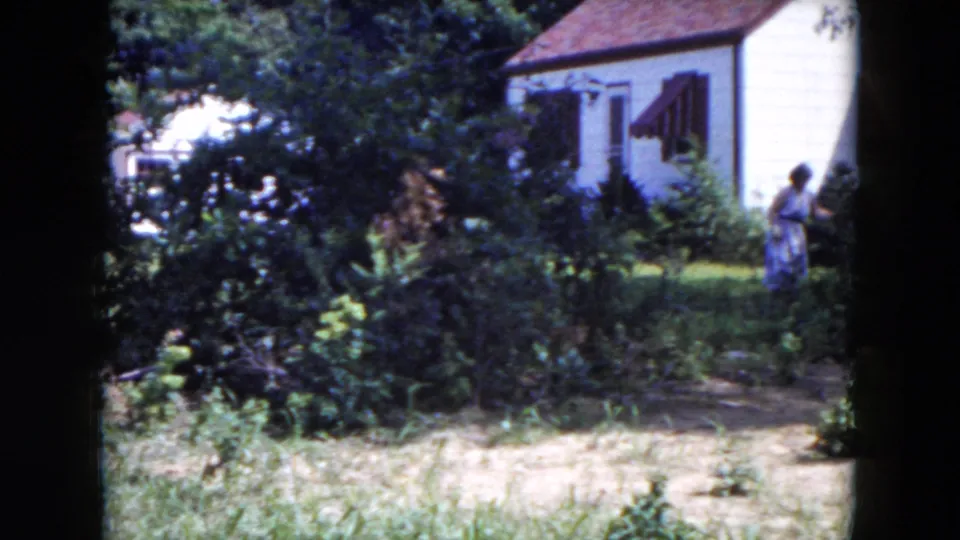 1954 Hicksville 8mm frame with a suburban house, garden growth, lawn, and a distant woman in a light dress near the yard.