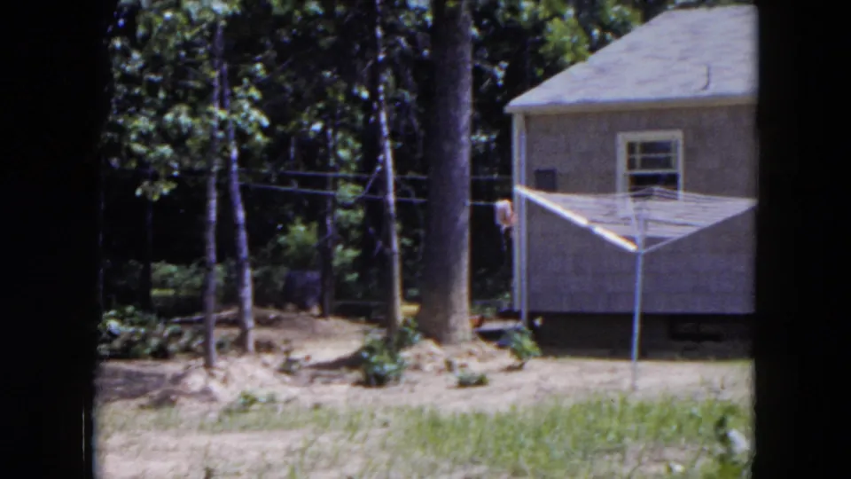 1954 Hicksville frame with a house exterior, wooded backdrop, patchy yard, overhead wires, and a white rotary clothesline.