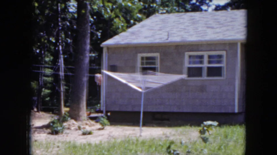 1954 Hicksville home-movie frame showing the side wall of a modest house, white windows, a clothesline, trees, and patchy lawn.