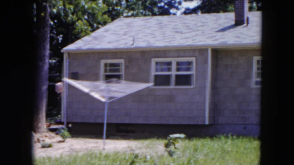 1954 Hicksville frame showing a modest house exterior with windows, roof, chimney, grass, trees, and a white rotary clothesline.