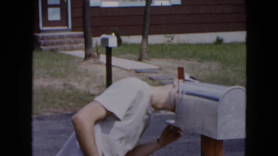 In Hicksville, New York, a man in a light T-shirt bends toward a gray curbside mailbox outside a suburban residence.