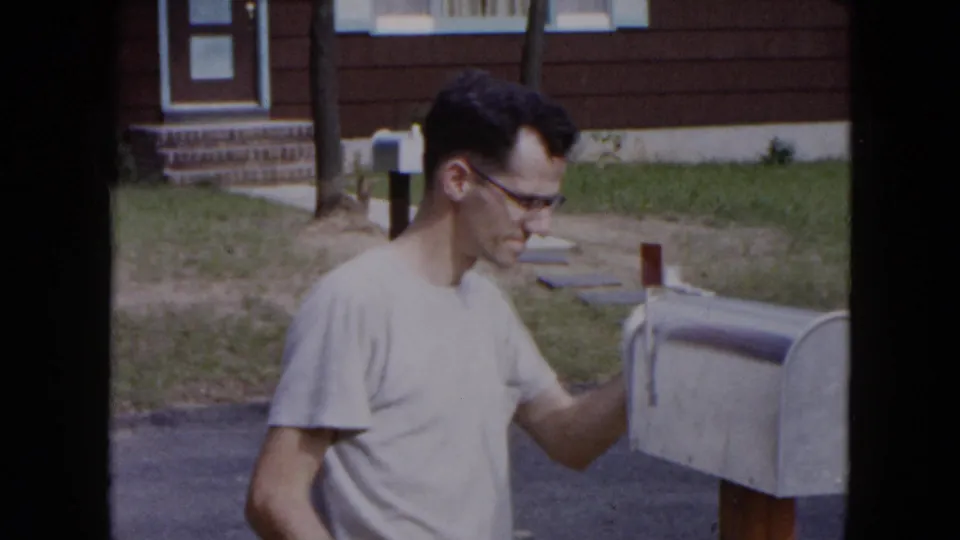 A man with glasses stands beside an open metal mailbox in a 1954 Hicksville suburban exterior, with lawn and house behind him.