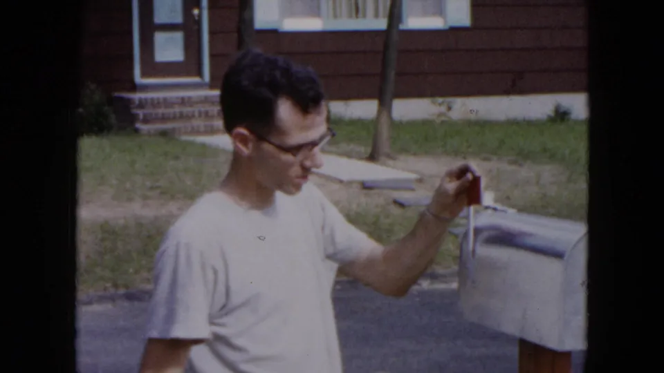 The man reaches toward the red flag on a gray curbside mailbox in front of a Hicksville residence, with lawn and steps visible.