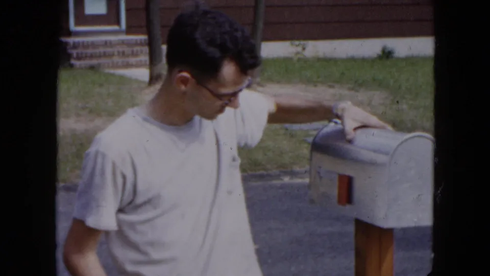 A man in glasses looks down while resting his hand on an open curbside mailbox outside a Hicksville suburban home.