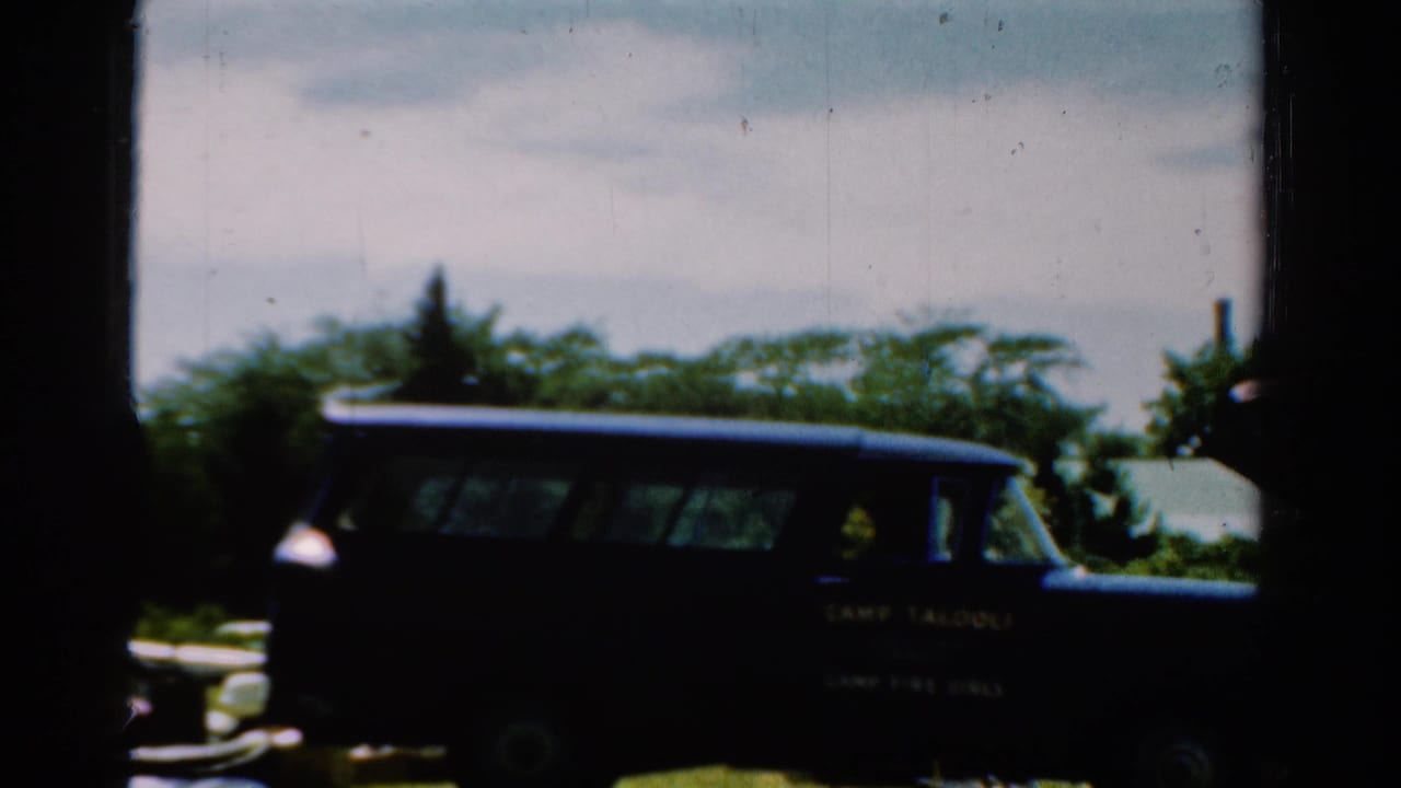 1954 California 8mm frame showing a dark camp-marked station wagon parked outdoors near trees and grass.