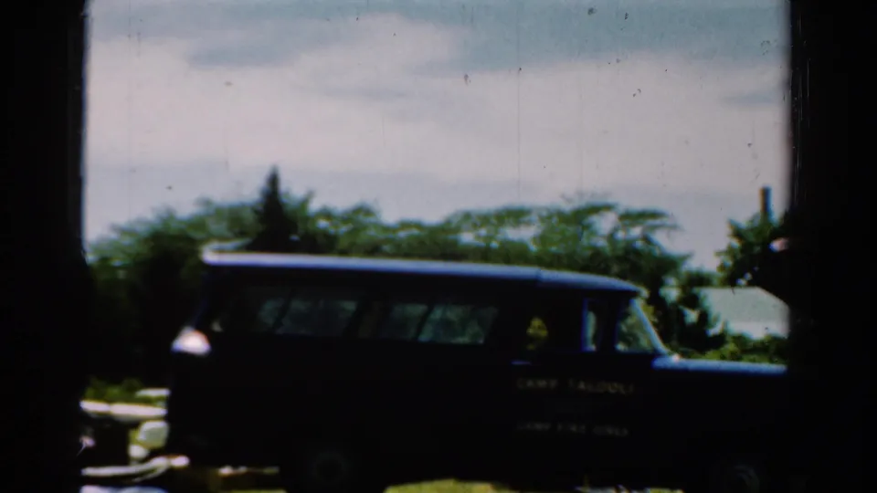 1954 California 8mm frame showing a dark camp-marked station wagon parked outdoors near trees and grass.
