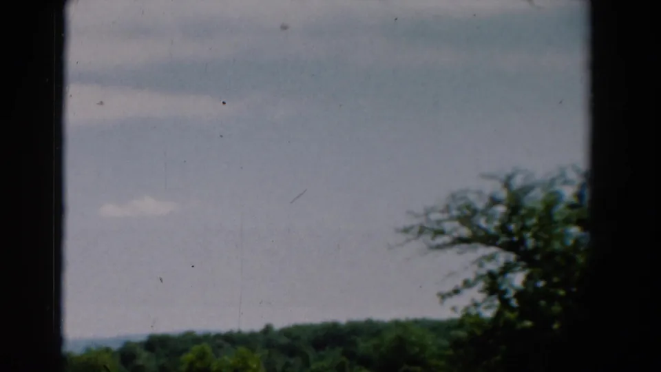 1954 California 8mm frame showing cloudy sky, a low wooded horizon, dark window framing, and foreground foliage.