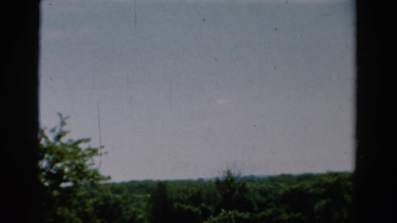 1954 California 8mm frame showing a soft view through dark vertical framing toward cloudy sky and a green treeline.