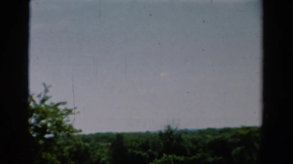 1954 California 8mm frame showing a soft view through dark vertical framing toward cloudy sky and a green treeline.