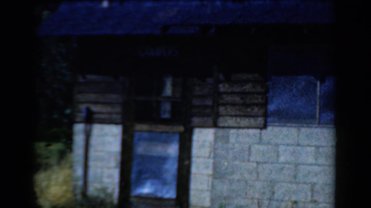 Rustic camp building facade with wood siding, concrete block base, small windows, and a sign above the entrance.