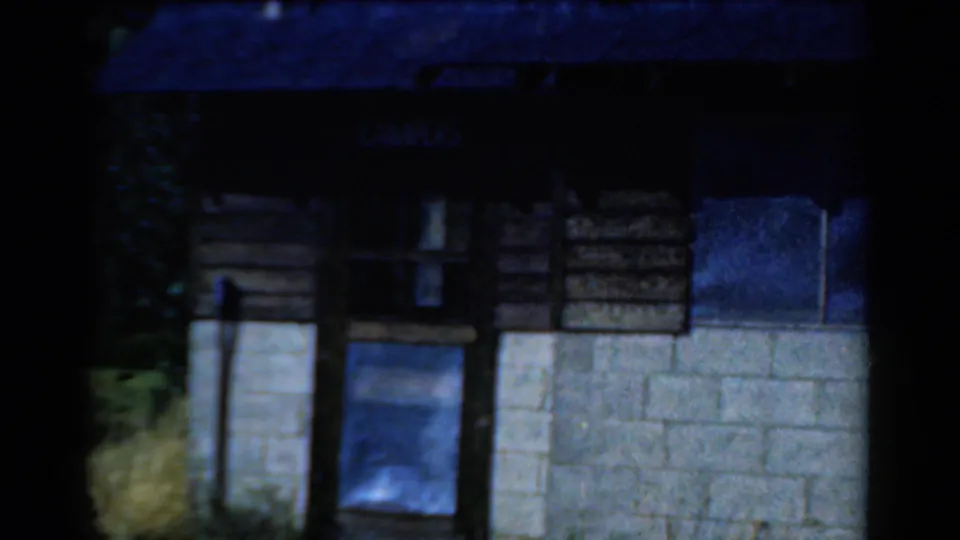 1954 California 8mm frame showing a rustic camp building exterior with wood siding, concrete blocks, windows, and a sign over the doorway.