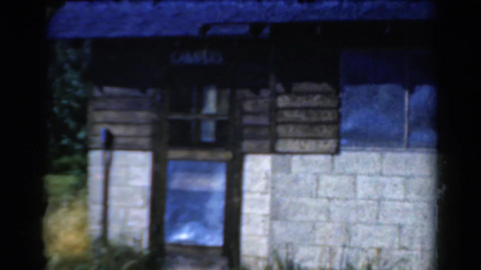 1954 California 8mm frame of a camp building front, with a readable camp sign, small window, door area, wood siding, and concrete block wall.