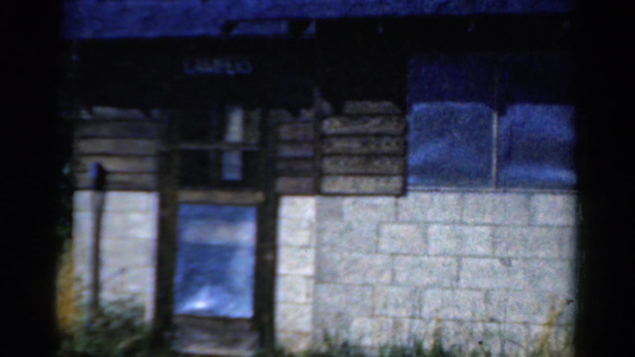 1954 California 8mm frame holding on a rustic camp building entrance area with wood siding, a small window, block foundation, and weeds below.