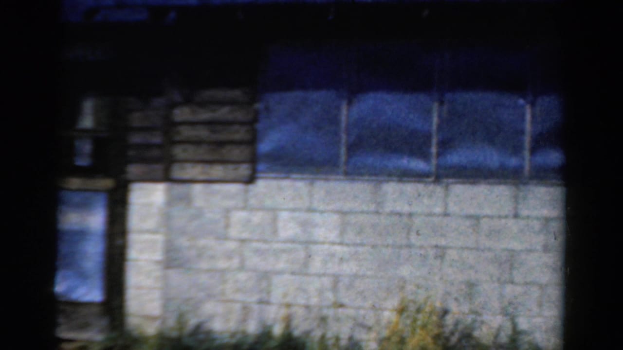 1954 California 8mm frame showing the side of a camp building with concrete block wall, wooden upper siding, windows, weeds, and heavy shadow.