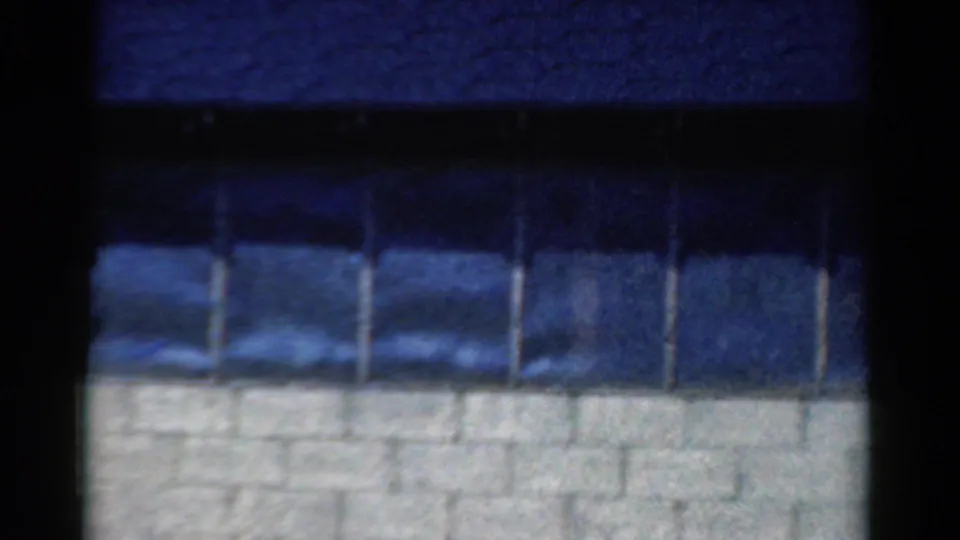 1954 California home-movie frame of blue-tinted window panels over a pale masonry wall, with visible grain and dark borders.
