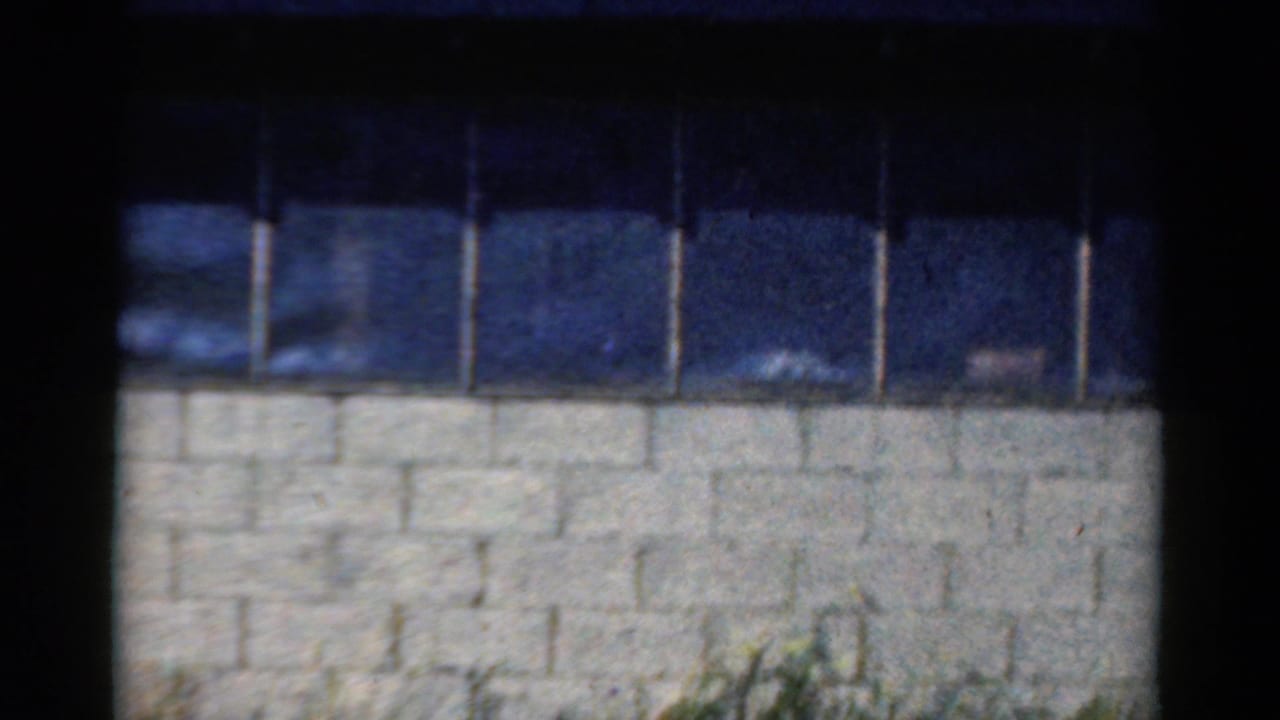 1954 California 8mm frame showing dark blue windows, a white cinder block wall, black borders, and faint grass at the bottom.