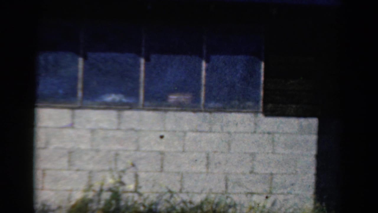 1954 California home-movie frame of a blue window band above a white block wall, with dark film borders and a shadowed right edge.