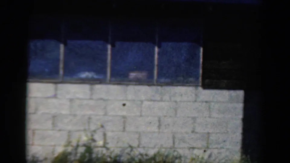 1954 California home-movie frame of a blue window band above a white block wall, with dark film borders and a shadowed right edge.