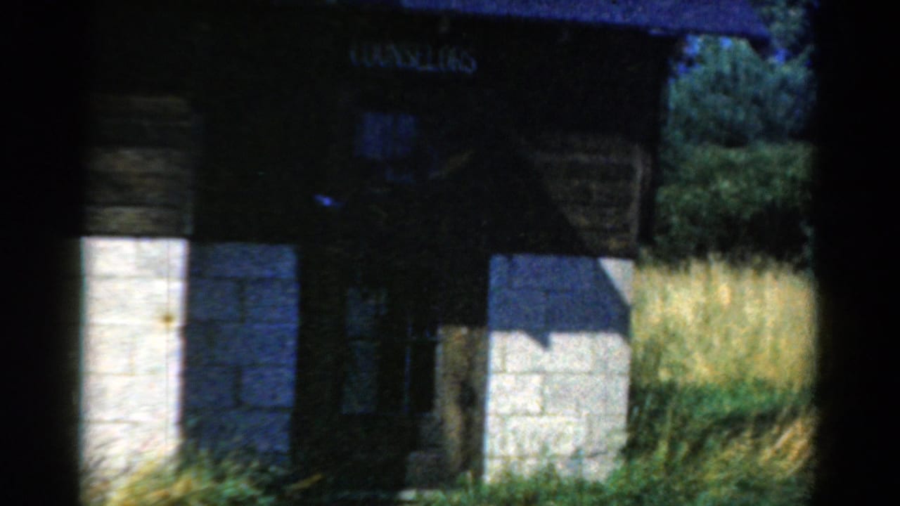 1954 California 8mm frame showing the COUNSELORS sign above a shadowed camp-building entrance with tall grass nearby.