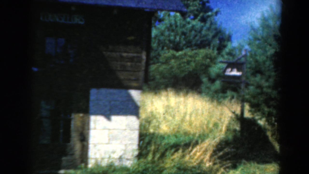 1954 California 8mm frame showing the COUNSELORS building partly in shadow, with tall grasses, trees, blue sky, and a small signpost.