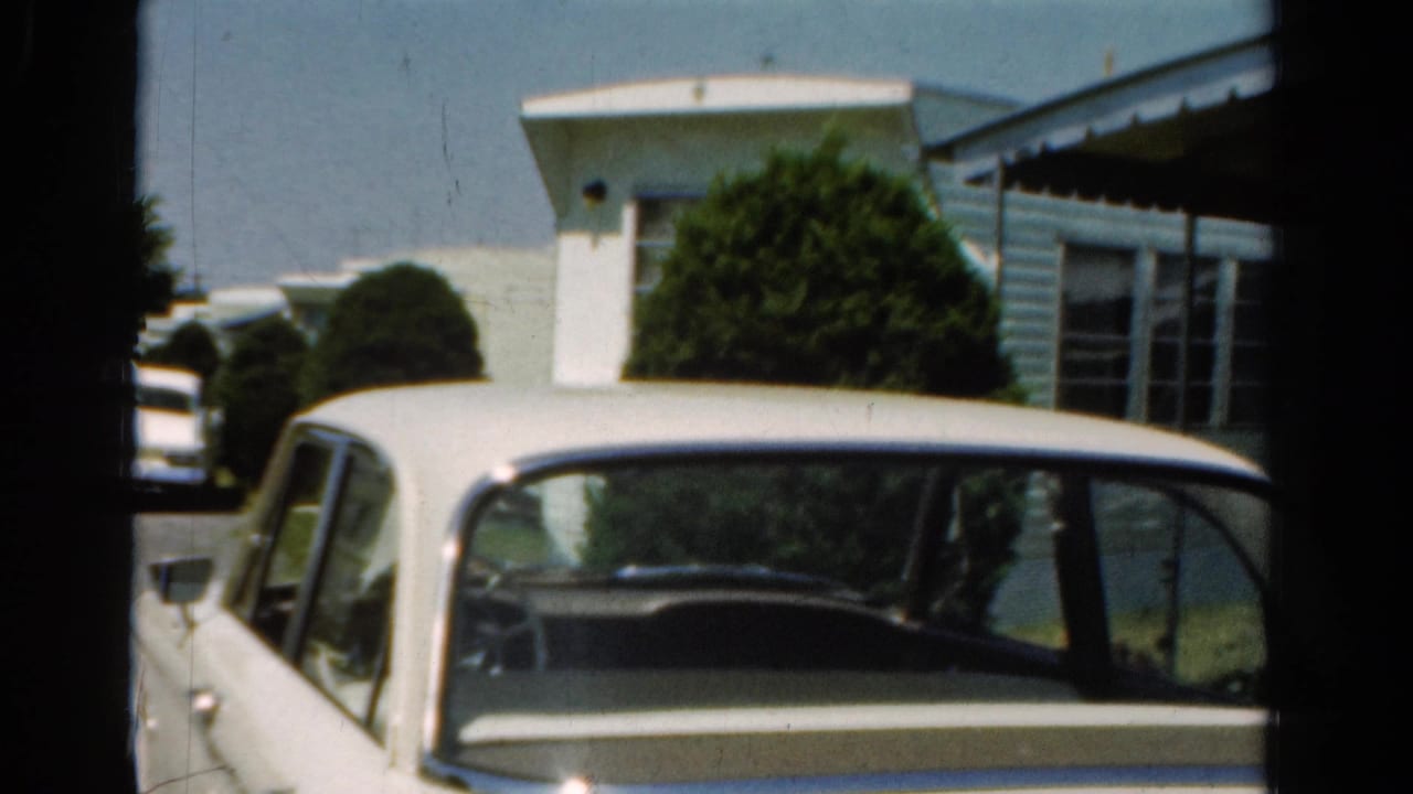 1954 California home-movie frame of a white mid-century sedan parked near a light mobile home or prefabricated house.