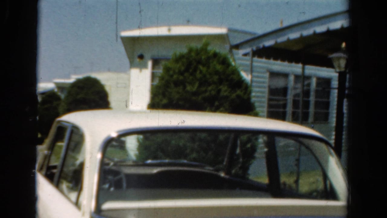 Close 8mm frame of a white mid-century sedan partly cropped below a light mobile home and green shrubbery.