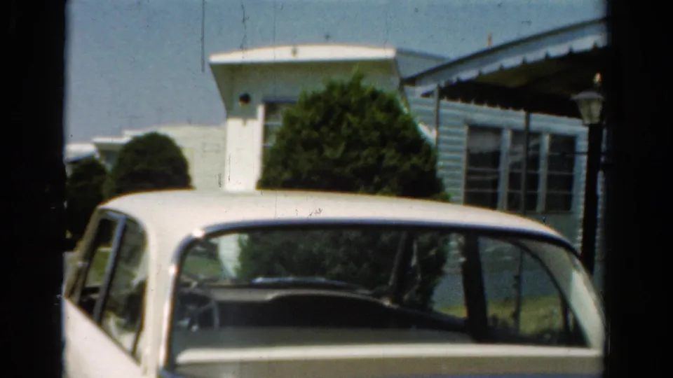 Close 8mm frame of a white mid-century sedan partly cropped below a light mobile home and green shrubbery.
