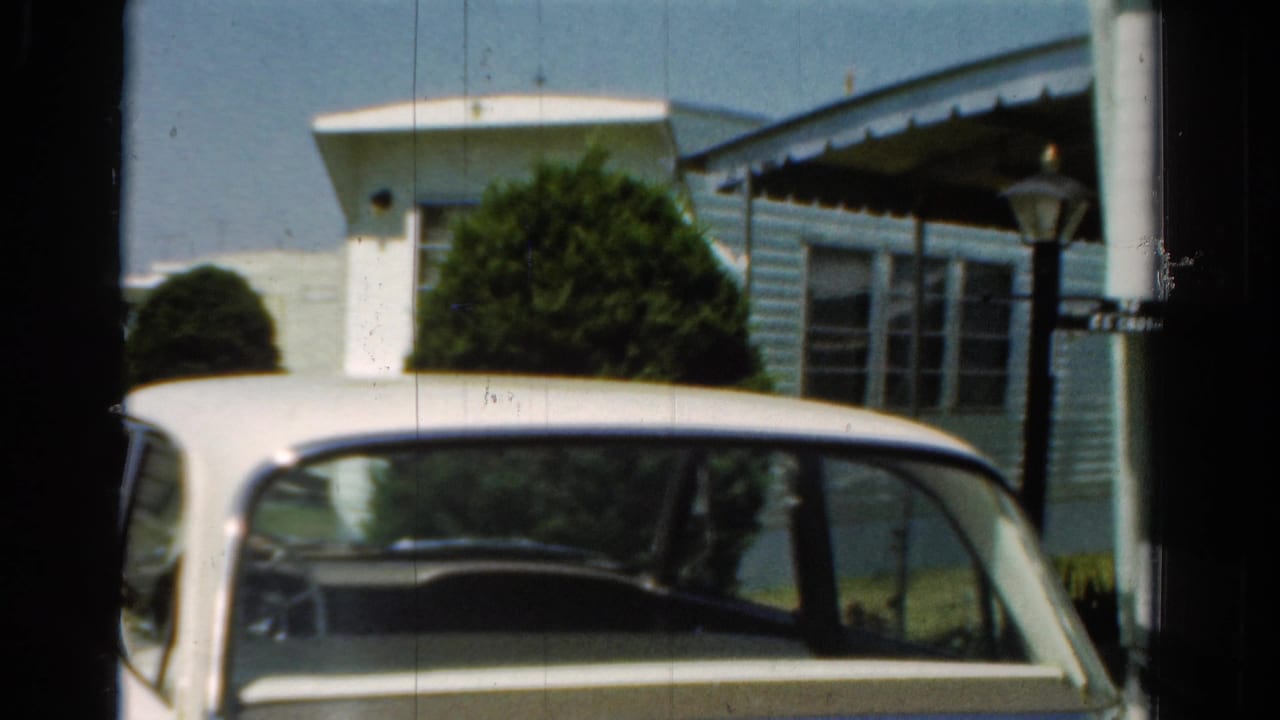 White car roof and windshield in front of a light blue house exterior with shrubs and a porch.
