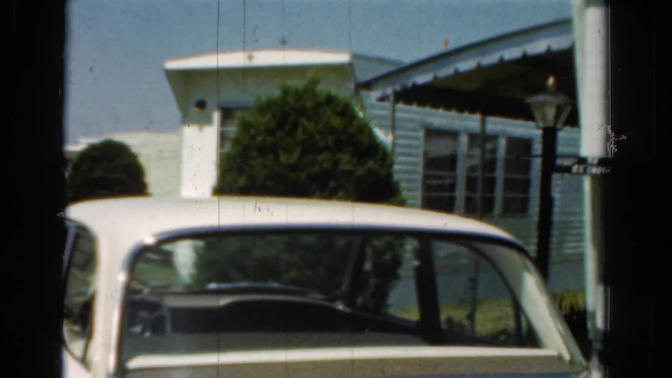1954 California 8mm frame showing a white car foreground and a small light blue residential exterior with shrubs, porch awning, and lamp post.