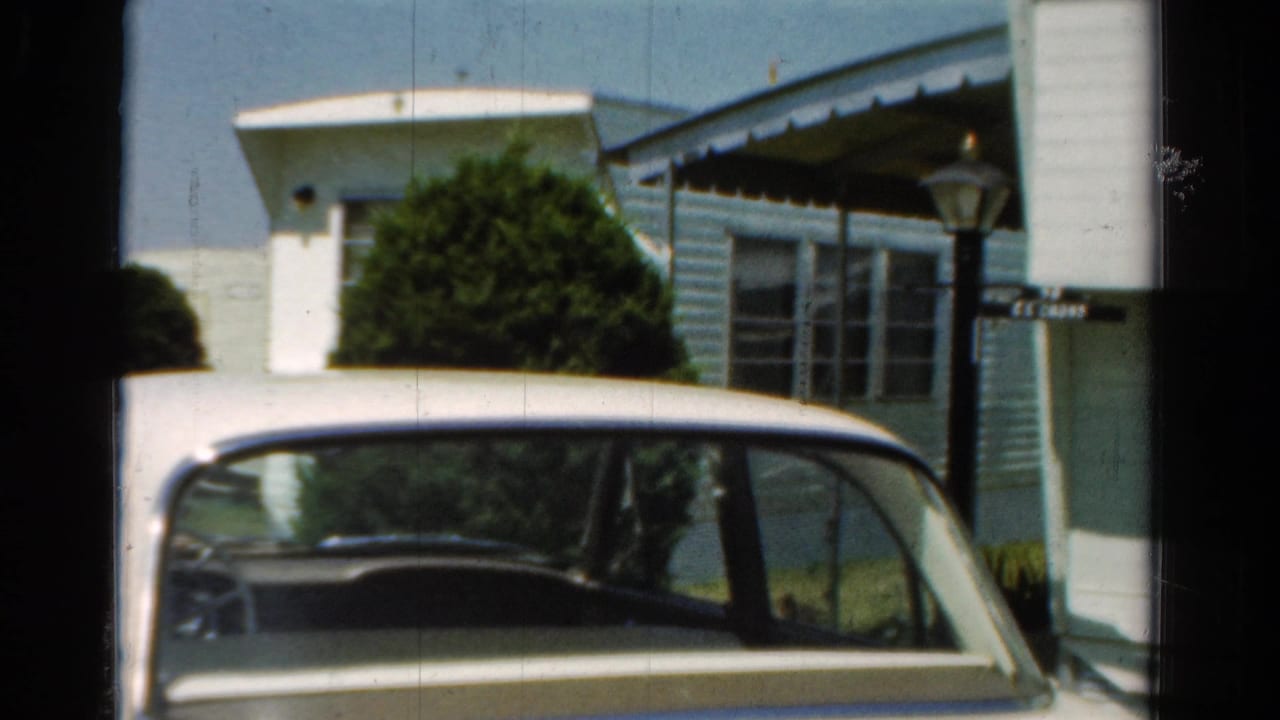 Parked white car beside a light blue building, shrubs, a lamp post, and a small sign.