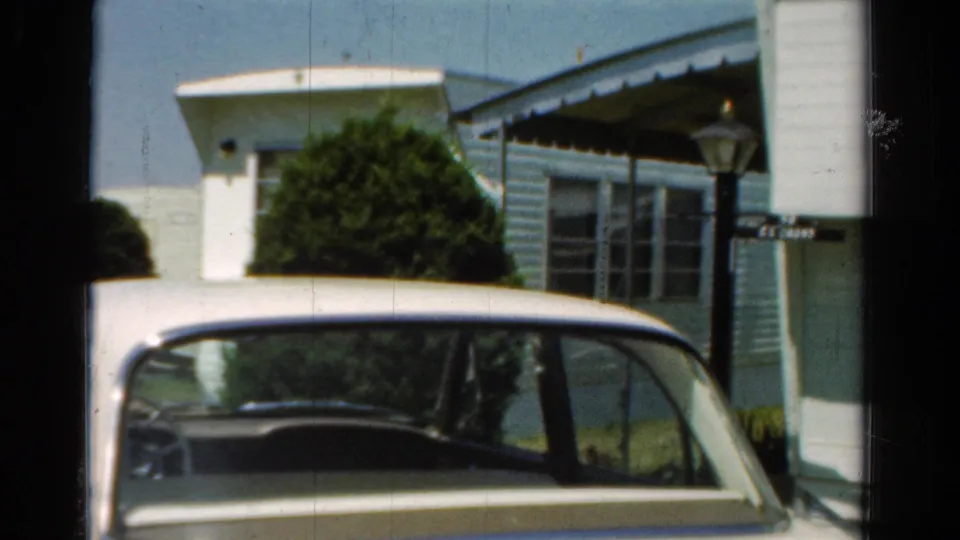 1954 California home-movie frame with a parked white car, pale blue house or mobile-home exterior, porch awning, shrubs, lamp post, and small sign.