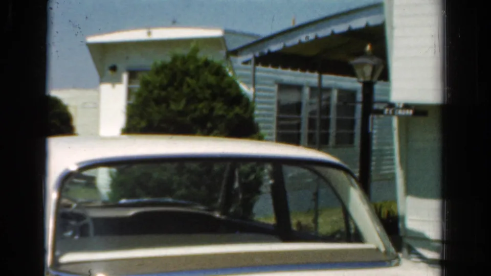 1954 California 8mm view of a white car in the foreground and a sunlit blue residential exterior with shrubs and a covered porch.