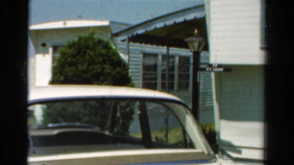 Final 1954 California frame showing the parked white car, light blue house or mobile-home exterior, porch awning, shrubbery, lamp post, and small sign.